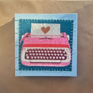 Puede incluir: Ilustración de una máquina de escribir rosa con un corazón marrón en una hoja de papel, sobre un fondo azul. La imagen está enmarcada por una tela azul claro con costuras blancas, creando una estética artesanal. La obra de arte está sobre un lienzo cuadrado.