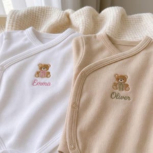 Könnte beinhalten: Zwei langärmelige Wickelbodys für Babys in Weiß und Beige. Beide sind mit einem gestickten braunen Teddybären mit Schleife sowie einem personalisierten Namen in Schreibschrift versehen: Emma in Rosa und Oliver in Salbeigrün.