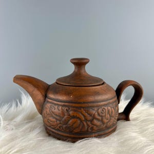 May include: A brown ceramic teapot with a lid and handle. The teapot has a floral relief design around the body. The spout and handle are curved. The teapot is on a white, fluffy surface.