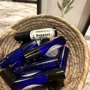 Könnte beinhalten: Ein geflochtener Korb gefüllt mit blauen Glas-Rollerflaschen mit schwarzen Kappen. Eine Flasche trägt die Aufschrift "Immune Support Roller". Der Korb steht auf einer Marmoroberfläche, im Hintergrund gerahmte botanische Drucke.