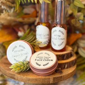 May include: A wooden cake stand displays beauty products, including two glass bottles with silver caps and labels reading "Pure Glow" and "Facial Toner." Two small, round tins are labeled "Exfoliating Sugar Scrub" and "Whipped Tallow Honey Butter Face Cream."