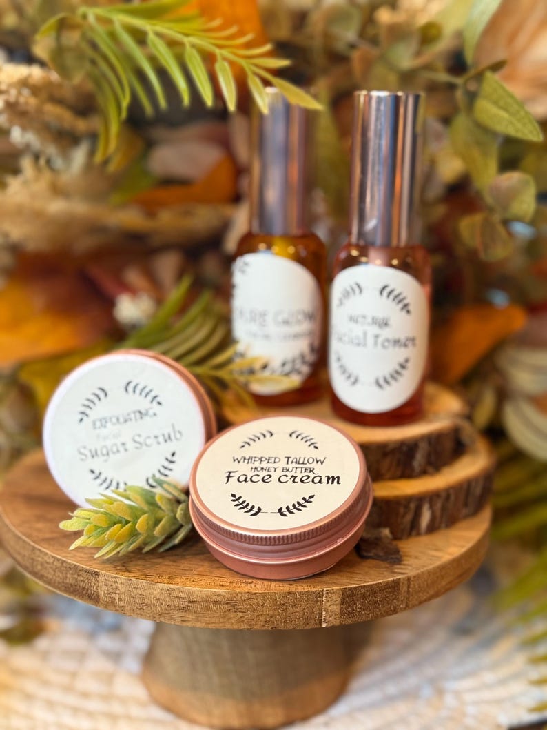 May include: A wooden cake stand displays beauty products, including two glass bottles with silver caps and labels reading "Pure Glow" and "Facial Toner." Two small, round tins are labeled "Exfoliating Sugar Scrub" and "Whipped Tallow Honey Butter Face Cream."