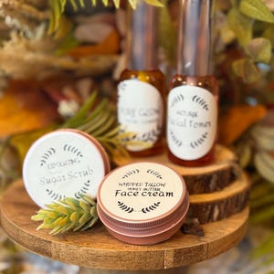 May include: A wooden pedestal displays skincare products. Two glass bottles with silver caps and labels reading "Pure Glow" and "Facial Toner" are visible. Two small, round tins labeled "Exfoliating Sugar Scrub" and "Whipped Tallow Honey Butter Face Cream" are also present.