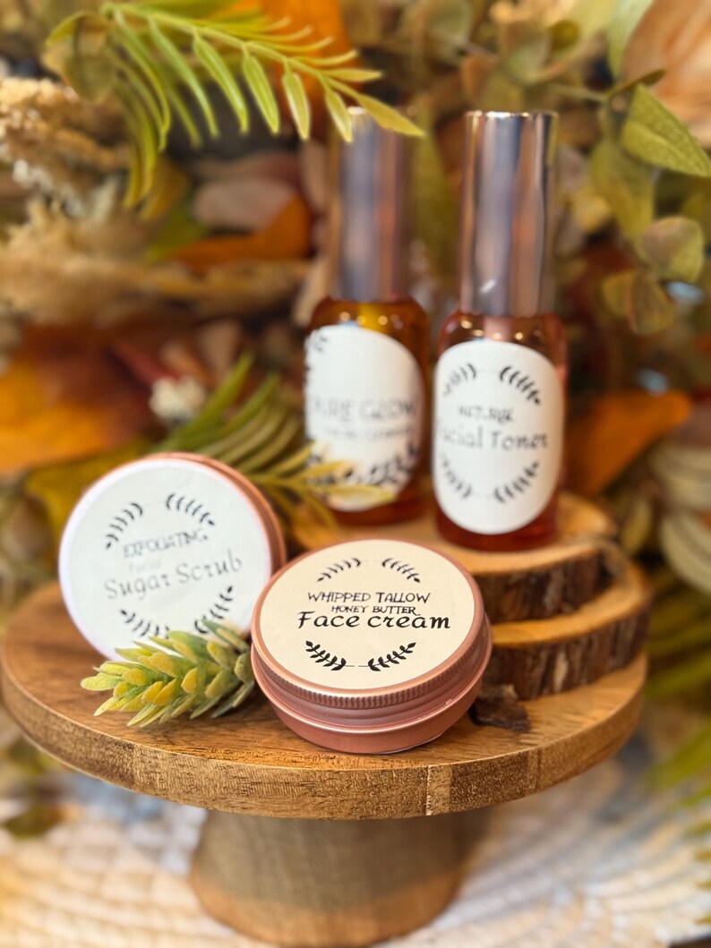 May include: A wooden pedestal displays skincare products. Two glass bottles with silver caps and labels reading "Pure Glow" and "Facial Toner" are visible. Two small, round tins labeled "Exfoliating Sugar Scrub" and "Whipped Tallow Honey Butter Face Cream" are also present.