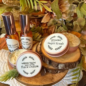 May include: A collection of skincare products, including facial cleanser, toner, sugar scrub, and face cream, displayed on a wooden tray. The products are in small bottles and tins with labels. The background features autumn-themed foliage.