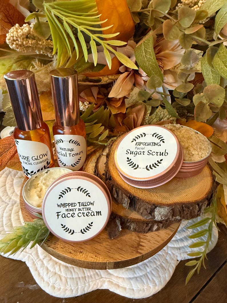 May include: A collection of skincare products, including facial cleanser, toner, sugar scrub, and face cream, displayed on a wooden tray. The products are in small bottles and tins with labels. The background features autumn-themed foliage.
