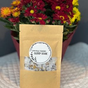 Peut inclure: Un sac en papier brun avec une fenêtre transparente contenant un produit de bain relaxant. Le sac porte une étiquette ronde blanche avec le texte "LAVENDER & CHAMOMILE SLEEP SOAK". Un bouquet de fleurs rouges et jaunes est en arrière-plan.