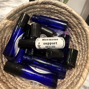 Könnte beinhalten: Ein geflochtener Korb gefüllt mit mehreren kobaltblauen Rollerflaschen mit schwarzen Kappen. Eine Flasche trägt die Aufschrift "Hormone support ROLLER". Die Flaschen sind in einem Korb aus Naturfasern angeordnet.
