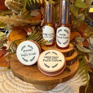 May include: A display of skincare products, including two glass bottles with silver tops labeled "Pure Glow" and "Natural Facial Toner." Two round containers are labeled "Exfoliating Facial Sugar Scrub" and "Whipped Tallow Honey Butter Face Cream."