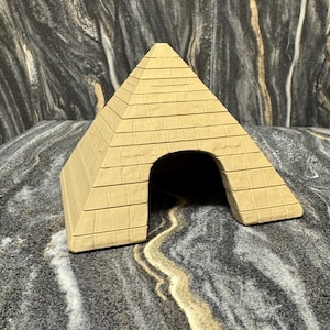 Puede incluir: Una pequeña pirámide de color beige, con una base rectangular y una parte superior triangular. La pirámide tiene una sola abertura arqueada y líneas horizontales que representan los bloques de piedra. El fondo es un patrón oscuro y veteado.