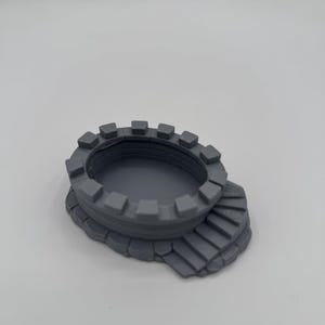 Può includere: Un pezzo di terreno in miniatura stampato in 3D di colore grigio, caratterizzato da una struttura circolare a muro di pietra merlata con un centro cavo. La base include una serie di gradini in pietra stampati che portano alla cima del muro.