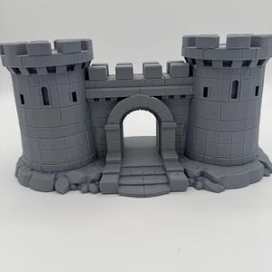 Può includere: Un modello di porta di castello stampato in 3D di colore grigio, caratterizzato da due torri cilindriche con merlature, un ingresso centrale ad arco e una base con texture in pietra. La struttura presenta un motivo a mattoni modulari.