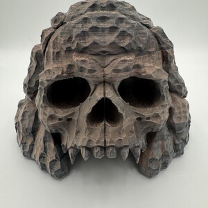Può includere: Una scultura dettagliata di teschio grigio con un aspetto strutturato e scolpito. Il teschio presenta grandi orbite oculari, una prominente cavità nasale e denti affilati. La parte superiore del teschio ha un design ruvido e stratificato, che gli conferisce un aspetto invecchiato.