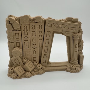 Può includere: Un pezzo di terreno in miniatura color marrone chiaro che raffigura un muro di pietra in rovina con una porta ad arco. La struttura presenta simboli geroglifici scolpiti, una colonna sul lato destro e detriti sparsi, tra cui blocchi rettangolari e piccoli teschi.