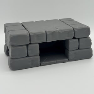 Op de afbeelding: Een grijze, rechthoekige, steenachtige structuur met een holle opening in het midden. Het object is opgebouwd uit meerdere blokvormige segmenten met gladde, afgeronde randen, die doen denken aan een miniatuurmuur of een kleine grot.