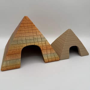 Può includere: Due sculture a forma di piramide in miniatura. La piramide più grande è una miscela di tonalità arancioni, gialle e verdi, con un motivo a mattoni strutturato. La piramide più piccola è di colore beige uniforme. Entrambe hanno aperture ad arco.