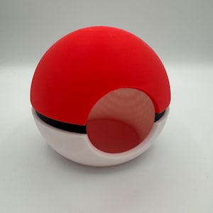Puede incluir: Objeto rojo y blanco con forma de Poké Ball con una franja negra. La mitad superior es roja, la mitad inferior es blanca y hay una abertura circular en el lateral. Probablemente un artículo decorativo o un contenedor.