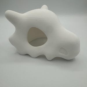 Peut inclure: Sculpture blanche en forme de crâne de Cubone. La sculpture présente une grande orbite oculaire et une petite protubérance en forme de corne. Fabriqué dans un matériau mat et texturé.