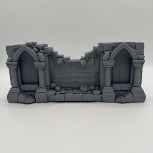Peut inclure: Une pièce de terrain miniature grise représentant un mur en pierre en ruine avec deux alcôves voûtées. La structure présente des briques détaillées, des bords effrités et des décombres éparpillés à la base pour les jeux de plateau.