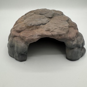 Può includere: Una struttura a forma di grotta strutturata, nei toni del grigio e del marrone. La formazione rocciosa artificiale ha un'apertura ad arco e una superficie ruvida e irregolare. Questo pezzo decorativo è probabilmente per un habitat di rettili o piccoli animali.