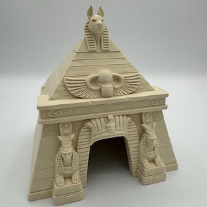 Op de afbeelding: Een beige, miniatuur Egyptische piramidevormige structuur. De piramide heeft gedetailleerde gravures van een Anubis-kop, een scarabee met vleugels en farao-beelden. De ingang is een gewelfde doorgang.