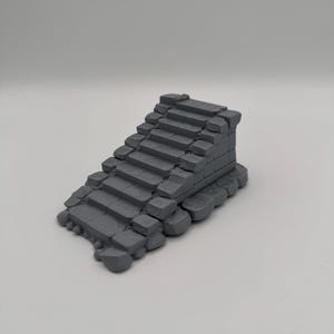Pode incluir: Um modelo de escada em miniatura impresso em 3D na cor cinza com textura de pedra desgastada. A estrutura consiste em vários degraus retangulares com bordas irregulares e ásperas, projetada para se assemelhar a um conjunto de ruínas antigas.