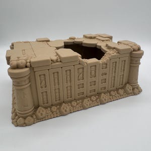 Könnte beinhalten: Ein hellbeiger, rechteckiger Taschentuchbox-Überzug mit einem detaillierten architektonischen Design, vertikalen Säulen, gestapelten Steinstrukturen und einer zentralen Öffnung. Die Basis ist von kleinen, abgerundeten, steinartigen Dekorelementen umgeben.