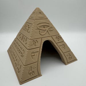 Può includere: Una piramide color beige, stampata in 3D, con incisioni geroglifiche e il simbolo dell'Occhio di Horus. La piramide ha un'apertura e presenta un design a tema antico Egitto. La struttura è realizzata in materiale solido.
