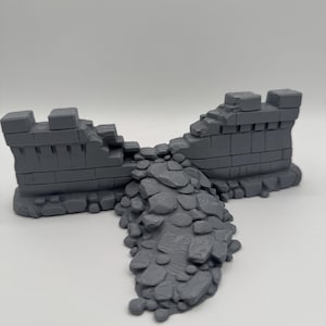 Op de afbeelding: Een grijs 3D-geprint miniatuur terreinstuk met een beschadigd stenen muurgedeelte met een centrale opening. Een hoop puin en losse stenen verspreidt zich vanuit de opening in de muur naar buiten, wat een getextureerd puinveld creëert.