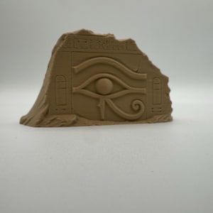 Può includere: Una scultura color sabbia simile alla pietra, a forma di frammento di muro irregolare. La superficie presenta un bassorilievo dell'Occhio di Horus, affiancato da due pannelli rettangolari verticali e sormontato da una fila di geroglifici.