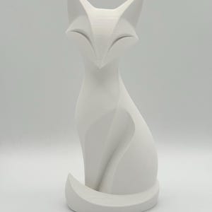 Könnte beinhalten: Eine matte, weiße Fuchsfigur mit einem stilisierten, geometrischen Design und scharfen, kantigen Linien. Der Fuchs ist in sitzender Position dargestellt, wobei sein Schwanz ordentlich um die Basis gewickelt ist und eine glatte Oberfläche zeigt.