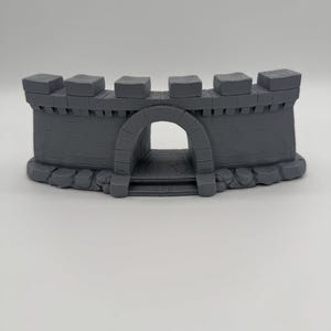 Pode incluir: Uma secção de muralha de castelo em miniatura cinzenta com um portão arqueado central, ameias ao longo do bordo superior e uma base de pedra texturada. A estrutura é moldada com linhas de tijolo horizontais e detalhes de pedra verticais.
