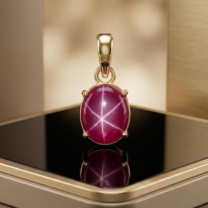 May include: A gold pendant featuring an oval-shaped, deep pink star ruby gemstone with a distinct six-rayed star pattern. The gemstone is held in a four-prong setting with a polished gold bail at the top, reflecting light on a smooth surface.