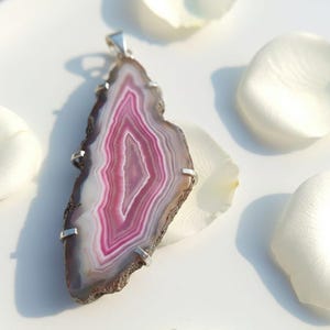 Pode incluir: Pingente de ágata polida com argola e detalhes prateados. A pedra apresenta faixas concêntricas em tons de branco, rosa e castanho. O pingente tem formato irregular, com borda natural. Pétalas de flores brancas no fundo.