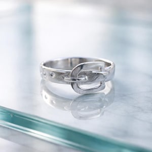 Könnte beinhalten: Ein silberner Ring mit einem Gürtelschnallen-Design. Der Ring hat eine detaillierte Schnalle mit strukturierter Oberfläche und einem kleinen Stift. Das Band hat kleine, gleichmäßig verteilte Löcher. Der Ring wird auf einer reflektierenden Oberfläche gezeigt.