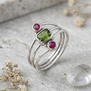 Peut inclure: Une bague en argent ornée d'une pierre précieuse verte rectangulaire centrale, entourée de deux petites pierres rouges rondes. L'anneau est composé de trois fils d'argent parallèles qui convergent vers une tige unique, créant un design ouvert.