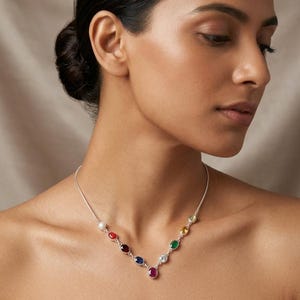 Puede incluir: Un collar de plata con una serie de piedras preciosas de colores en un diseño graduado. El collar incluye una perla, piedras rojas, moradas, azules y color rubí, junto con piedras verdes, amarillas y transparentes. El collar se lleva en el cuello de una persona.