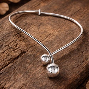 May include: A silver necklace with a distinctive design. It features a curved bar with two spherical accents. The necklace is presented on a wooden surface.