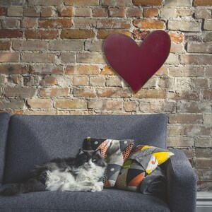 Puede incluir: Una decoraci&oacute;n de pared en forma de coraz&oacute;n burdeos cuelga sobre un sof&aacute; gris con cojines estampados. Un gato gris y blanco descansa en el sof&aacute;. El fondo es una pared de ladrillo con diferentes tonos de marr&oacute;n y rojo.
