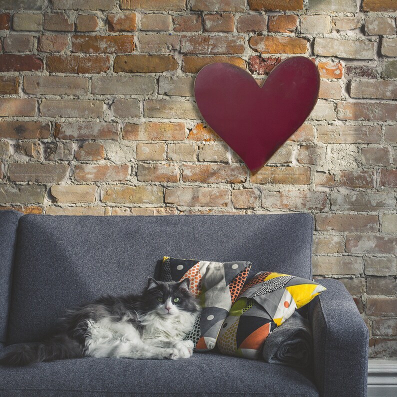 Puede incluir: Una decoraci&oacute;n de pared en forma de coraz&oacute;n burdeos cuelga sobre un sof&aacute; gris con cojines estampados. Un gato gris y blanco descansa en el sof&aacute;. El fondo es una pared de ladrillo con diferentes tonos de marr&oacute;n y rojo.