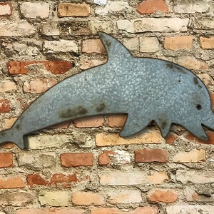 May include: A metal dolphin wall hanging with a distressed, galvanized finish. The dolphin is in a leaping pose, with a curved body and a smiling mouth. The wall hanging is mounted on a brick wall.