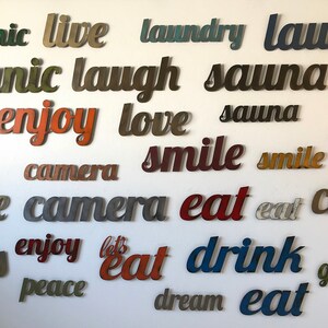 Puede incluir: Una colecci&oacute;n de recortes de palabras met&aacute;licas, incluyendo "enjoy", "eat", "smile" y "love", en varios colores como rojo, naranja y azul, montados en una pared blanca. Las palabras est&aacute;n en una fuente cursiva, creando una pieza de arte mural decorativa.