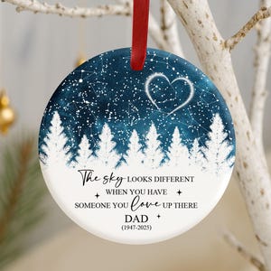 May include: A round Christmas ornament with a dark blue sky design, white stars, and a heart-shaped constellation. White trees line the bottom. Text reads "The sky looks different when you have someone you love up there, Dad (1947-2025)." A red ribbon is attached.