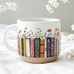 May include: White ceramic mug with a wraparound design of colorful books and floral accents. The books are in various colors, and the flowers include pink, yellow, and orange blooms. The mug has a tan base and a white handle.