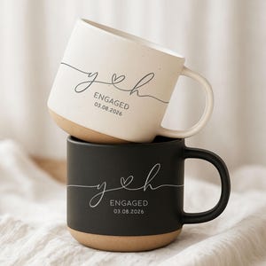May include: A stack of two ceramic mugs, one cream-colored with dark speckles and one matte black. Both feature the cursive letters y and h connected by a heart, with the text ENGAGED and the date 03.08.2026 printed in a clean, sans-serif font.
