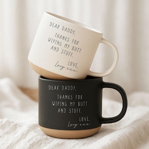 May include: Two ceramic mugs, one stacked on top of the other. The top mug is white with a tan base and handle, the bottom mug is black with a tan base and handle. Both mugs have the text "DEAR DADDY, THANKS FOR WIPING MY BUTT AND STUFF. LOVE, Lucy xxx."