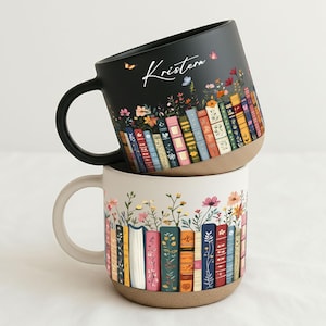 May include: Two ceramic mugs stacked on top of each other. The top mug is black with a floral and book design, and the name "Kristen" in white script. The bottom mug is cream-colored with a similar design. Both mugs have a brown base and a handle.