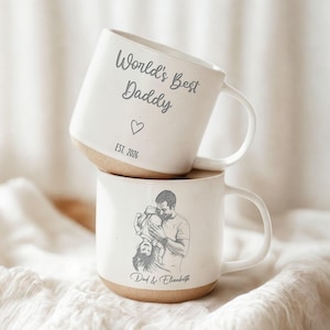 May include: Two white ceramic mugs with a tan base. The top mug reads "World's Best Daddy" with a heart and "EST. 2026". The bottom mug features a sketch of a father and child with the text "Dad & Elizabeth".