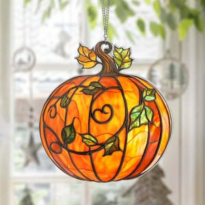 May include: Stained glass pumpkin decoration with orange and yellow hues, featuring a brown stem and green leaves. The pumpkin is suspended by a silver chain, perfect for fall or Halloween decor. The design includes intricate details and a classic seasonal theme.