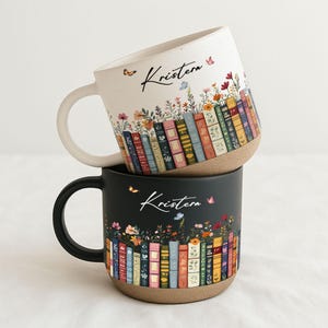 May include: Two ceramic mugs stacked on top of each other. The top mug is white with a cream-colored handle, and the bottom mug is black with a black handle. Both mugs feature a floral and book design with the name "Kristen".
